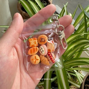 May include: A clear plastic pouch containing miniature food-shaped charms, including a square biscuit, a round bun, a croissant, and a hot dog. The pouch features a beige metal lobster clasp for attachment and the text HAPPY AT HOME CLUB.