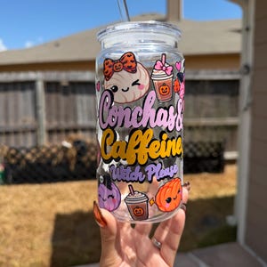 May include: A clear glass tumbler with a lid and straw, featuring colorful cartoon graphics of a concha, pumpkins, and coffee cups. The text reads Conchas & Caffeine Witch Please in pink, yellow, and purple lettering against the glass surface.