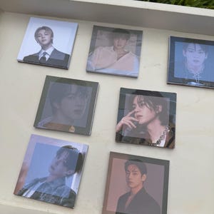 May include: A collection of seven square coasters featuring individual photographic portraits of seven different people. Each coaster displays a distinct headshot with varying color tones, including black and white, soft pink, and blue hues.