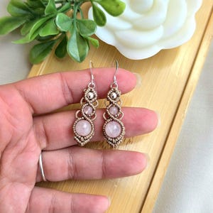 May include: A pair of dangle earrings featuring light brown macrame cord woven into intricate patterns, each holding a small silver-toned bead and two round, pale pink rose quartz stones. The earrings hang from silver-toned metal hooks.