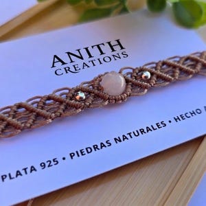 May include: A close-up of a handmade macrame choker necklace. The necklace is a light brown color with a central rose quartz stone and silver accents. The text "ANITH CREATIONS" is visible.