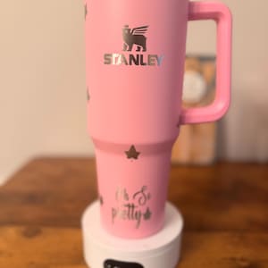 May include: A pink Stanley tumbler with a handle, featuring the Stanley logo and star accents. The text "Oh So pretty" is printed on the bottom. The tumbler is sitting on a white base.