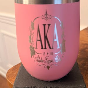 Può includere: Un bicchiere in acciaio inossidabile rosa con coperchio trasparente, caratterizzato da un design inciso color argento che include le lettere AKA, i numeri 1908 e il testo Alpha Kappa Alpha circondato da una cornice decorativa di edera.