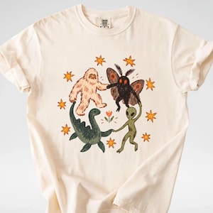 May include: A cream-colored short-sleeve t-shirt featuring a graphic print of a Bigfoot, a moth-like creature, a green aquatic dinosaur, and a gray alien holding hands in a circle, surrounded by orange stars and a small central flower.