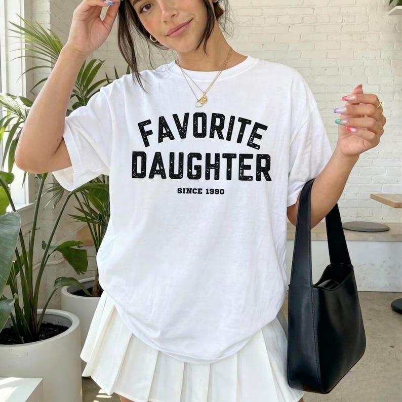 May include: White t-shirt with the text "FAVORITE DAUGHTER SINCE 1990" in black block letters. The shirt is worn with a white pleated skirt and a black shoulder bag. The image is taken in natural light.