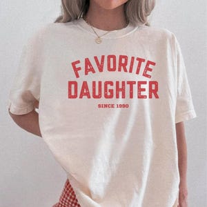 May include: Cream-colored t-shirt with the words "FAVORITE DAUGHTER" in red, distressed lettering. Below the text is "SINCE 1990" in a smaller red font. The shirt is a casual style, suitable for everyday wear.