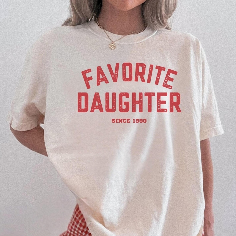 May include: Cream-colored t-shirt with the words "FAVORITE DAUGHTER" in red, distressed lettering. Below the text is "SINCE 1990" in a smaller red font. The shirt is a casual style, suitable for everyday wear.