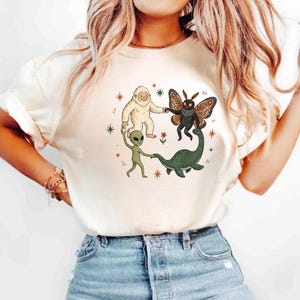 May include: A cream-coloured short-sleeve t-shirt featuring a central graphic illustration of a white yeti, a black moth creature, a green alien, and a green sea monster holding hands in a circle, surrounded by small orange and green star icons.