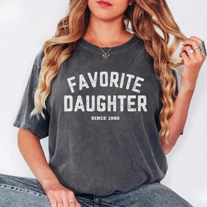 May include: Dark gray t-shirt with the text "FAVORITE DAUGHTER" in white, with "SINCE 1990" below. The shirt has a crew neck and short sleeves. The fabric appears soft and worn.