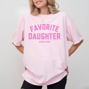 May include: A light pink t-shirt with the words "FAVORITE DAUGHTER" in distressed pink lettering. Below the text is "SINCE 1990". The shirt is a relaxed fit and has short sleeves.