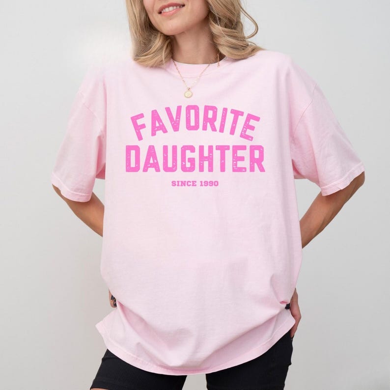 May include: A light pink t-shirt with the words "FAVORITE DAUGHTER" in distressed pink lettering. Below the text is "SINCE 1990". The shirt is a relaxed fit and has short sleeves.