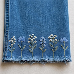 May include: A blue denim trouser leg featuring a frayed hem and a row of embroidered wildflowers along the bottom edge. The embroidery includes white daisies, blue cornflowers, and green stems, creating a detailed botanical pattern on the fabric.