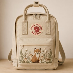 May include: A beige backpack featuring a circular Fjällräven Kånken logo patch at the top. The front pocket displays detailed embroidery of a fox sitting among white flowers, green ferns, and brown mushrooms. It has two top handles and zippers.