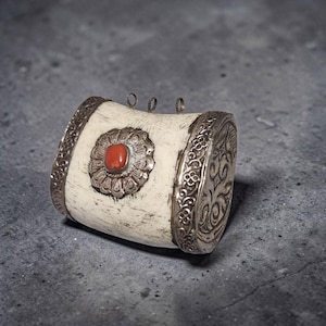 May include: A cylindrical pendant featuring a cream-colored bone body with ornate silver end caps and a central silver floral setting holding a smooth, oval-shaped orange stone. Three small silver loops are attached to the top edge of the piece.