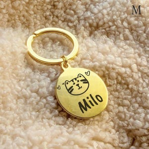 Pode incluir: Um porta-chaves dourado com um pingente redondo com uma ilustração de gato e o nome "Milo" em preto. O porta-chaves inclui um anel dividido e um pequeno laço. Dois pequenos corações são desenhados no pingente.