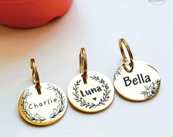 Op de afbeelding: Drie ronde goudkleurige metalen huisdierpenningen met ringen. Elke penning heeft een zwart gegraveerd bloemenkransontwerp en een naam: Charlie, Luna of Bella. De Luna-penning bevat een klein hartje. De tekst Photos of real products is zichtbaar.