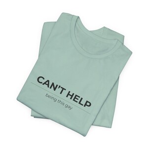 May include: A light green t-shirt with the text "CAN'T HELP being this gay" printed in black. The shirt is folded and has a crew neck.