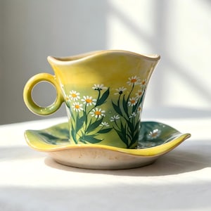 Peut inclure: Un ensemble tasse et soucoupe en céramique. La tasse et la soucoupe sont peintes avec un dégradé jaune et vert, et présentent un motif floral de marguerites. La tasse a une anse arrondie et un bord ondulé. La soucoupe est de forme assortie.