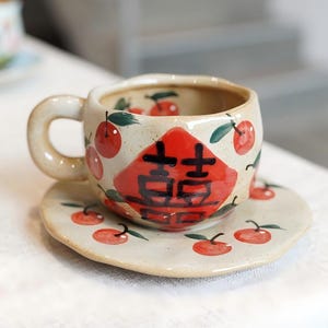 Peut inclure: Un ensemble tasse et soucoupe en céramique crème, orné d'illustrations de cerises rouges et de feuilles vertes. La tasse présente un grand emblème rouge avec des caractères noirs. L'ensemble est sur une surface blanche.