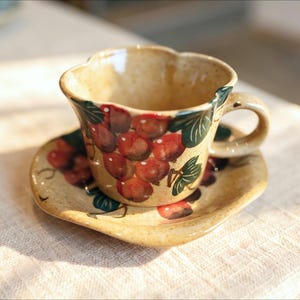 Peut inclure: Un ensemble tasse et soucoupe en céramique beige avec un bord festonné. La tasse et la soucoupe présentent un motif peint à la main de grappes de raisins rouges avec des feuilles vertes. La céramique a une finition mouchetée et un vernis brillant.