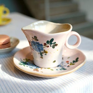 Może przedstawiać: Różowy ceramiczny zestaw filiżanki i spodka z kwiatowymi wzorami. Filiżanka ma falisty brzeg i mały uchwyt. Jasnobrązowy makaronik leży na małym talerzu. Kolejna żółta filiżanka jest w tle.