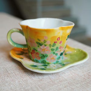 Peut inclure: Un ensemble tasse et soucoupe en céramique peinte à la main. La tasse et la soucoupe sont décorées de fleurs roses, de feuilles vertes et d'un fond jaune et vert. La tasse a un bord festonné et une anse verte.