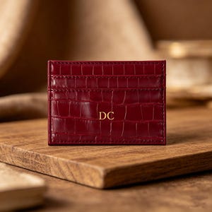 May include: A burgundy leather card holder with a crocodile texture. The card holder has multiple slots and the initials "DC" in gold lettering. The card holder is displayed on a wooden surface.