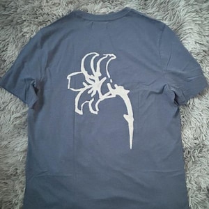 Peut inclure: Un t-shirt bleu ardoise à manches courtes orné d'un grand motif blanc dessiné à la main représentant une fleur de lys unique centrée au dos. Le vêtement présente une encolure ronde et une coupe droite simple avec des manches courtes.