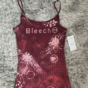 Peut inclure: Un débardeur à fines bretelles bordeaux avec un imprimé graphique rose clair portant le texte Bleech 9:3. Le vêtement présente des motifs circulaires abstraits, des éclaboussures et une étiquette blanche indiquant la taille 2XS.