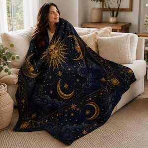 May include: A dark blue throw blanket featuring a gold celestial pattern with a central sun face, crescent moons, and scattered stars. The design includes swirling cloud motifs and decorative borders, creating a detailed night sky theme.