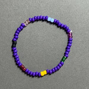 May include: A beaded bracelet made of small, round, purple beads. It has a variety of colourful accent beads, including black, red, yellow, green, pink, and light blue. The bracelet is strung on an elastic band.