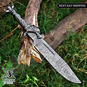 May include: A fixed-blade knife with a patterned Damascus steel blade and a dark metallic handle featuring an intricate dragon head guard and pommel. The blade displays a distinct wavy layered texture, and the handle has a textured grip.