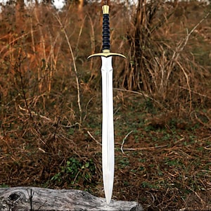 May include: A double-edged sword featuring a polished silver-toned steel blade, a gold-toned crossguard, and a black ridged grip with a gold-toned pommel. The sword is positioned vertically, showcasing its symmetrical design and pointed tip.
