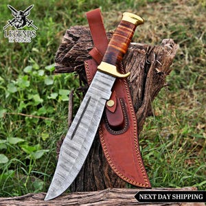 May include: A fixed-blade knife with a patterned Damascus steel blade, a stacked leather handle, and a brass pommel and guard. The knife is accompanied by a brown leather sheath featuring decorative stitching and a brass snap button closure.