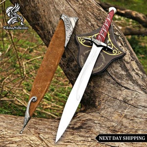 May include: A silver-toned metal sword with a red handle featuring white swirling patterns and a pointed blade. The sword is accompanied by a brown suede scabbard with silver metal accents at the tip and hilt, resting on a black display plaque.