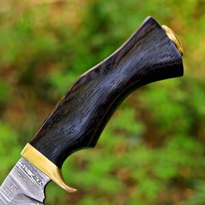 May include: A knife handle crafted from dark, textured wood with a prominent grain pattern. The handle features a polished brass bolster and a brass pommel cap at the end, connecting to a blade with a visible layered Damascus steel pattern.