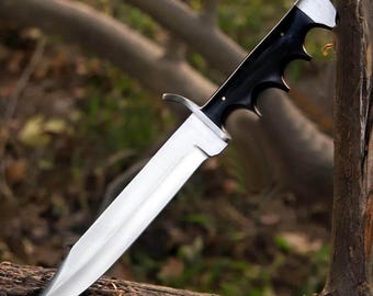 May include: A fixed-blade knife featuring a polished silver-toned steel blade with a prominent central ridge and a sharp point. The handle is crafted from smooth black material with a contoured finger-grooved grip and a silver-toned metal guard.
