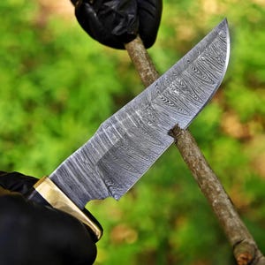 May include: A fixed-blade knife featuring a distinctive Damascus steel blade with a complex, wavy, layered pattern in shades of silver and dark gray. The knife includes a polished brass bolster and a dark handle, shown cutting into a small wooden branch.