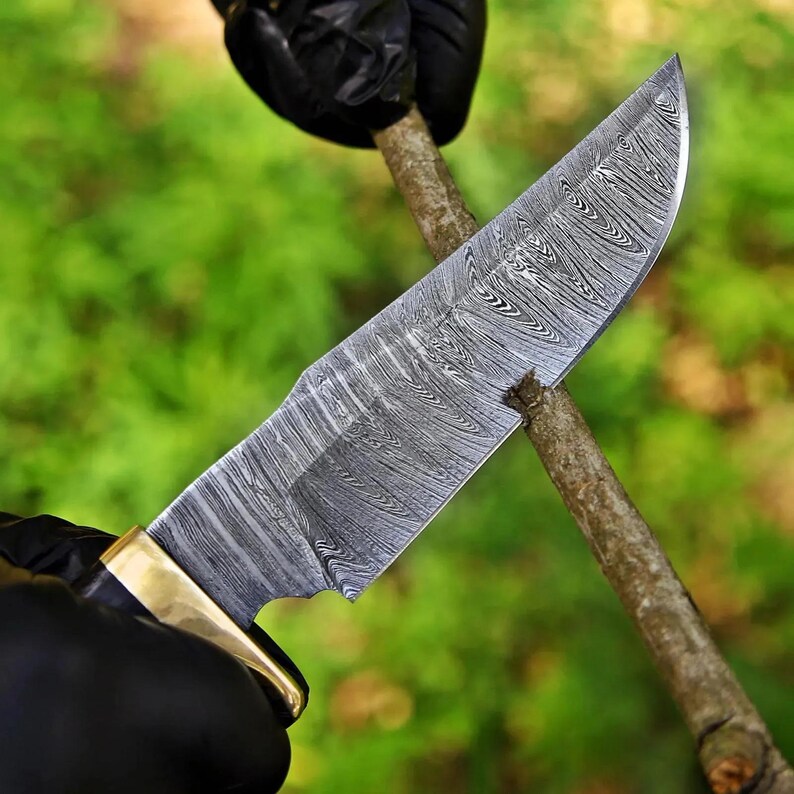 May include: A fixed-blade knife featuring a distinctive Damascus steel blade with a complex, wavy, layered pattern in shades of silver and dark gray. The knife includes a polished brass bolster and a dark handle, shown cutting into a small wooden branch.
