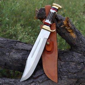 May include: A fixed-blade knife with a polished steel blade featuring a central fuller. The handle is crafted from textured antler with brass pommel and guard, accented by red, white, and blue rings. It includes a tan leather sheath with stitching.