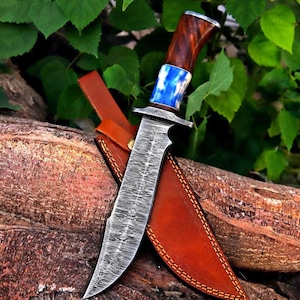 May include: A fixed-blade knife with a patterned Damascus steel blade, a silver-coloured bolster, a blue and white marbled handle section, and a dark wood grip. The knife is accompanied by a tan leather sheath with visible stitching along the edges.