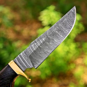 May include: A fixed-blade knife featuring a sharp, pointed steel blade with a distinct, swirling gray and black Damascus pattern. The knife includes a polished gold-colored metal bolster and a dark, textured handle with visible wood grain.