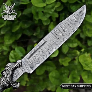 May include: A fixed-blade knife featuring a silver-toned metal handle shaped like a wolf head and a sharp blade with a distinct wavy Damascus steel pattern. The blade includes a central fuller groove. The text Legends of Vikings is visible.
