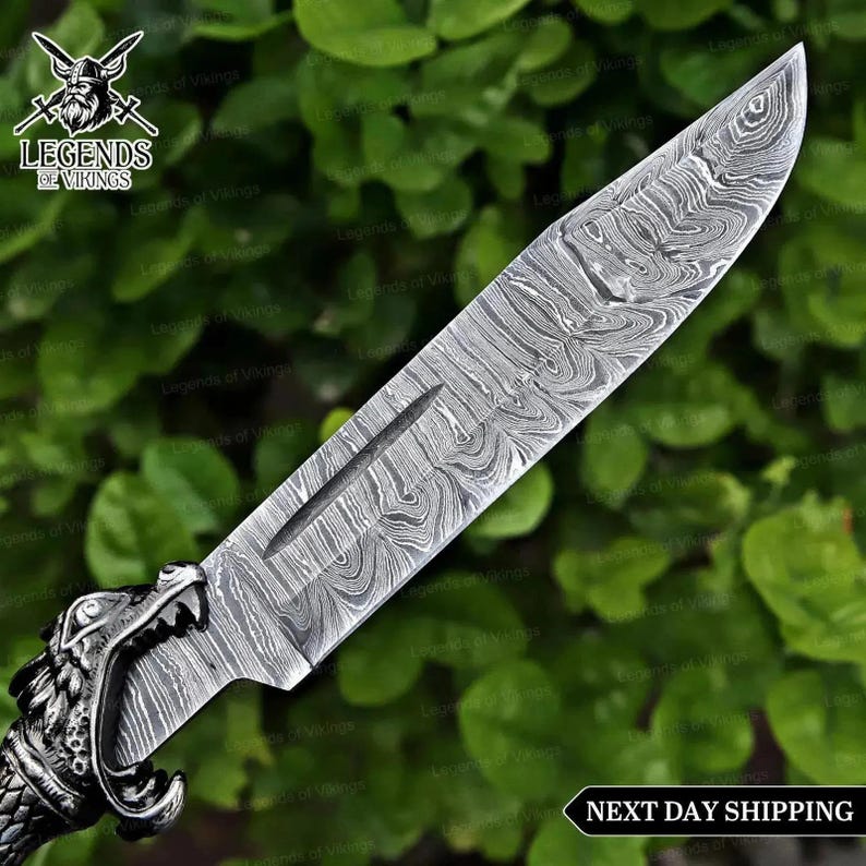 May include: A fixed-blade knife featuring a silver-toned metal handle shaped like a wolf head and a sharp blade with a distinct wavy Damascus steel pattern. The blade includes a central fuller groove. The text Legends of Vikings is visible.