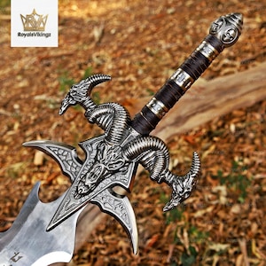Puede incluir: Una espada decorativa con una empuñadura de metal plateado y un diseño de cráneo de carnero en la cruz. El mango está envuelto en material oscuro con detalles metálicos y el pomo tiene forma de esfera con varios grabados de calaveras.