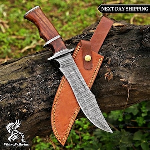 May include: A fixed-blade knife with a patterned Damascus steel blade and a polished dark wood handle featuring silver-toned metal bolsters. The knife is accompanied by a tan leather sheath with visible stitching and a brass-coloured snap button.