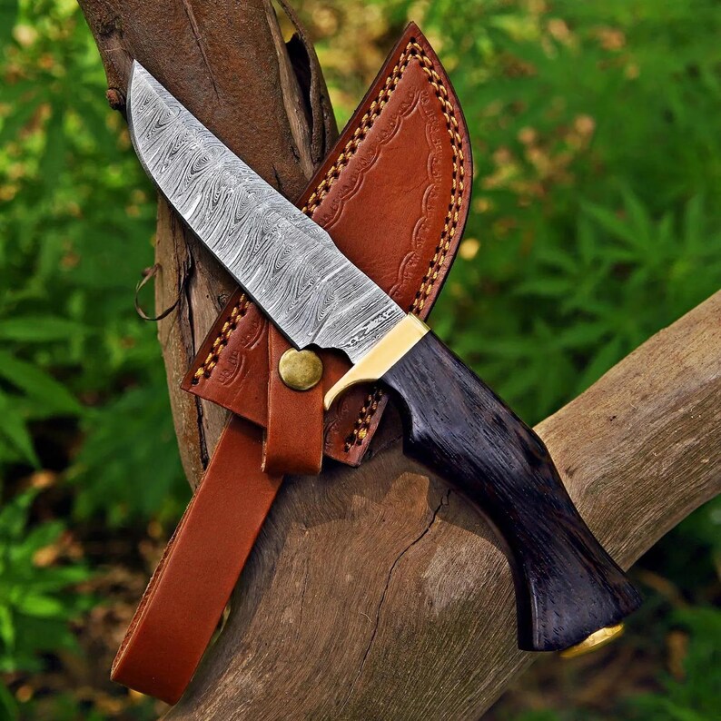 May include: A fixed-blade knife featuring a patterned Damascus steel blade, a dark wood handle with a brass bolster and pommel, and a tan leather sheath with decorative embossed edges and a single brass snap button closure.