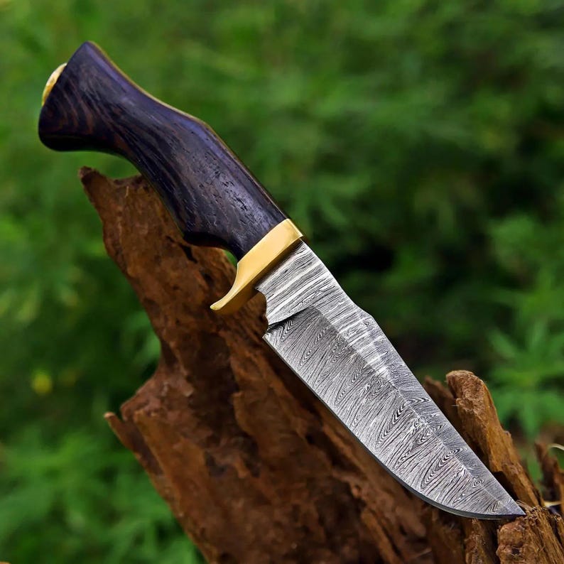 May include: A fixed-blade knife featuring a patterned Damascus steel blade with a distinct wavy texture. The handle is crafted from dark, polished wood with a visible grain, accented by a gold-colored metal bolster and a matching pommel cap.