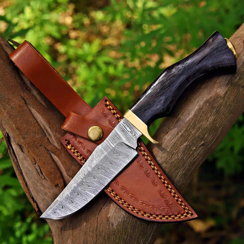May include: A fixed-blade knife featuring a patterned Damascus steel blade, a dark wood handle with a brass bolster, and a tan leather sheath with decorative stitching and a brass snap closure, designed for outdoor utility and carry.
