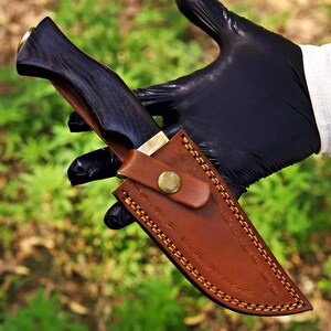 May include: A fixed-blade knife with a dark, polished wooden handle and a brass bolster, encased in a tan leather sheath. The sheath features decorative embossed patterns along the edges, yellow contrast stitching, and a brass snap button closure.